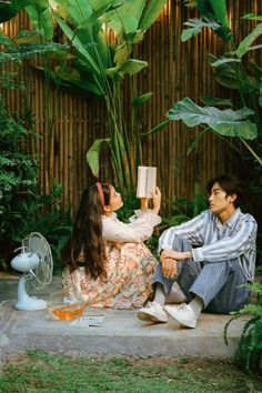 a man and woman sitting on the ground in front of a bamboo tree reading a book