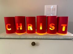 three red candles that spell out the word christ on a white surface with an electrical outlet in the background