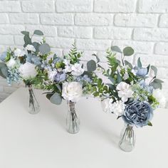 three vases with flowers and greenery are on a white table next to a brick wall