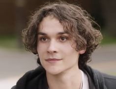 a young man with curly hair wearing a black jacket and white shirt is looking at the camera