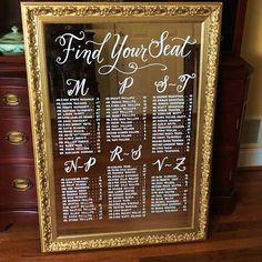 a wedding seating chart is displayed in a gold framed frame on a wooden floor next to a dresser