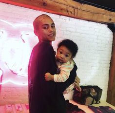 a man holding a baby in front of a white brick wall with neon lights on it