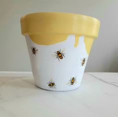 a yellow and white flower pot with bees painted on the side, sitting on a marble table