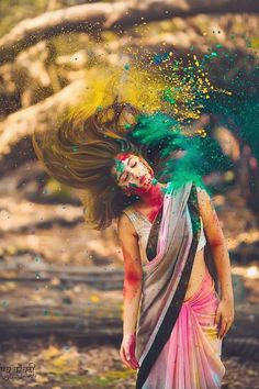 a woman with green and yellow paint on her face is standing in the woods while throwing colored powder into the air