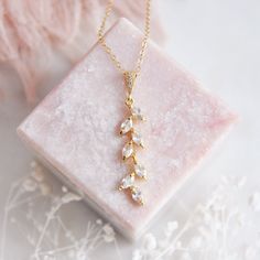 a gold necklace with white stones on a pink marble block next to some dried flowers