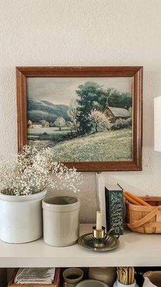 a white shelf topped with pots and baskets filled with flowers next to a painting on the wall