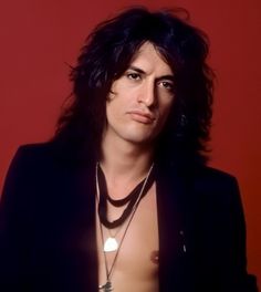 a man with long hair wearing a black jacket and silver necklace on his neck is looking at the camera