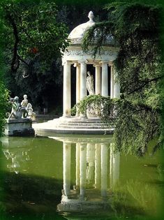 a gazebo in the middle of a pond surrounded by trees and statues with green foliage around it