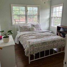 a bed sitting in a bedroom next to two windows and a book shelf on top of a hard wood floor