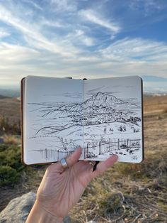 a hand holding up an open book in front of a mountain range and blue sky