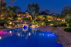 a large pool surrounded by trees and rocks with lights in the water at night time