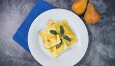 a white plate topped with ravioli next to two pears and a blue napkin