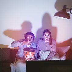 two people are sitting on a couch with popcorn in their hands and one is holding a remote control