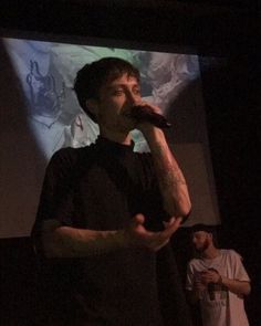 a man standing in front of a projection screen holding a microphone to his mouth while another man stands behind him