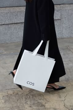 a woman walking down the street carrying a white shopping bag with cos written on it