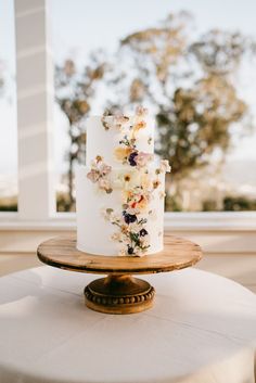 a white cake with flowers on it sitting on top of a wooden stand in front of a window