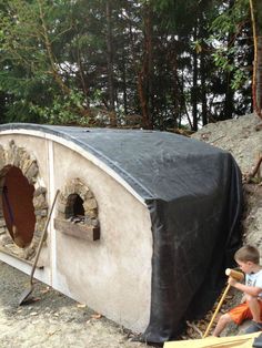 a young boy sitting on the ground next to an outdoor oven with a cover over it