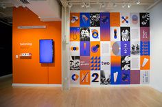 an orange and blue wall with posters on it in a museum like space that has wooden floors