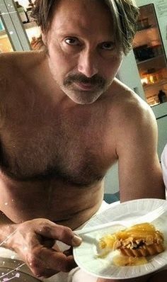 a shirtless man holding a plate with food on it and looking at the camera