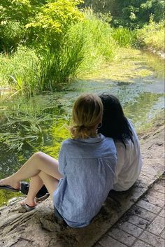 two women sitting on the edge of a river looking out at the grass and water