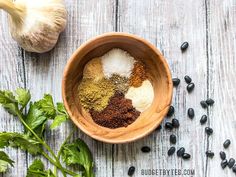 a wooden bowl filled with spices next to black beans and green leaves on top of a white wood table