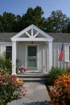 a small white house with an american flag on the front door and flowers around it