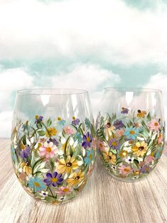 two hand painted wine glasses sitting on top of a wooden table with clouds in the background