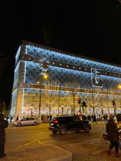 people are walking on the sidewalk in front of a building lit up with christmas lights