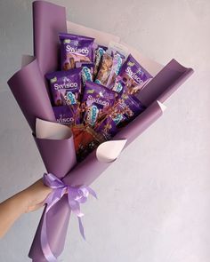 a hand holding a purple box filled with chocolates and wrapped in satin ribbon on top of a white wall