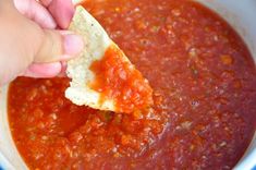 a hand holding a tortilla chip over a bowl of tomato sauce and cheese