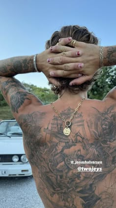 a man with tattoos covering his face and hands behind his head, in front of a car