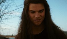 a man with long hair looking down at his cell phone while standing in front of a tree