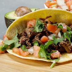 two tacos with meat and vegetables on a cutting board next to an avocado