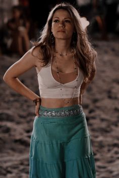 a woman standing in the sand with her hands on her hips wearing a white tank top and green skirt