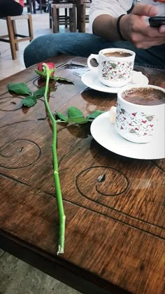 two cups of coffee sit on a table with a single rose in the foreground