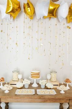 a table topped with cakes and desserts covered in gold foil balloon garlands hanging from the ceiling