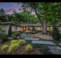 a house that is surrounded by trees and plants in front of the house at dusk