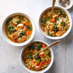 three bowls of chicken and vegetable soup on a table with spoons next to it