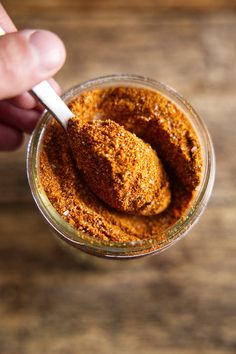 a hand holding a spoon over a small jar filled with spices on top of a wooden table