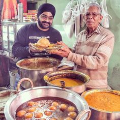 two men standing next to each other in front of pots filled with food