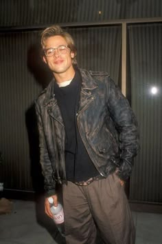 a man in glasses and a leather jacket is holding a bottle of alcohol while standing next to a wall