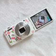 a white camera sitting on top of a bed next to a cell phone with a pink flowered design