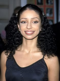 a woman with long black curly hair wearing a black dress and smiling at the camera