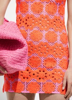 a woman in an orange and pink crochet dress holding a pink purse with holes on it