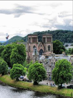 an old church is next to a river and some trees in the foreground with people walking on the other side