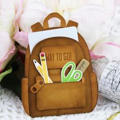 a back pack with scissors, pencils and markers in it next to some flowers