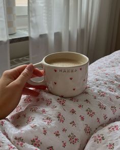 a hand holding a cup of coffee on top of a bed with white sheets and pink flowers