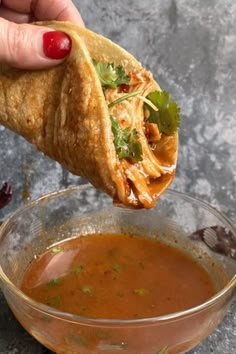 a hand holding a tortilla over a bowl of tomato soup and garnished with cilantro