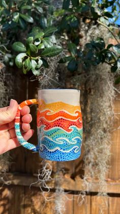 a hand holding a colorful coffee mug in front of a tree