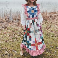 a woman in a dress and hat standing on the grass near water with her hands behind her back